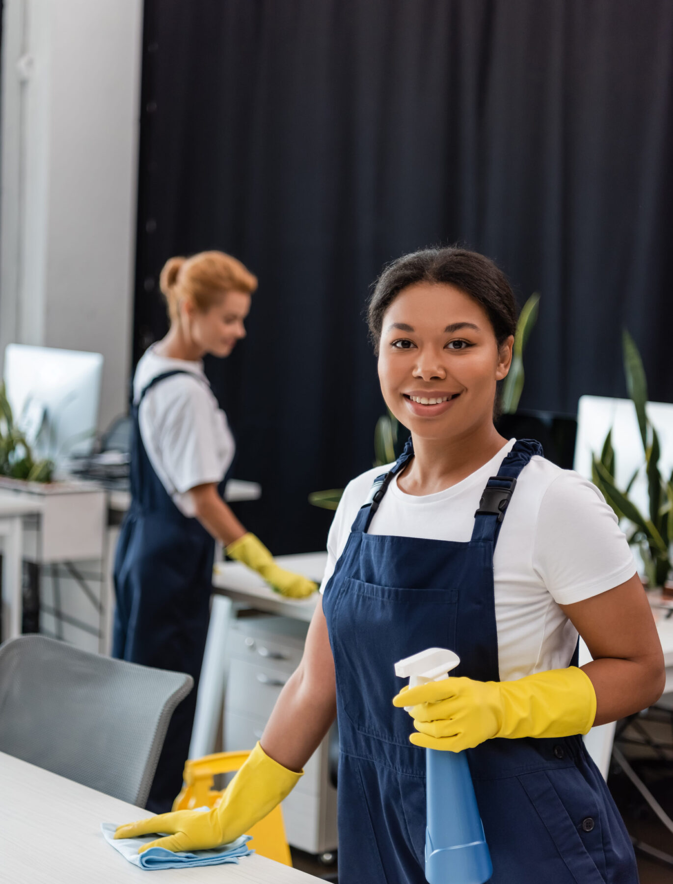 192264513_m happy bi-racial woman in overalls and rubber gloves smiling at camera near blurred colleague